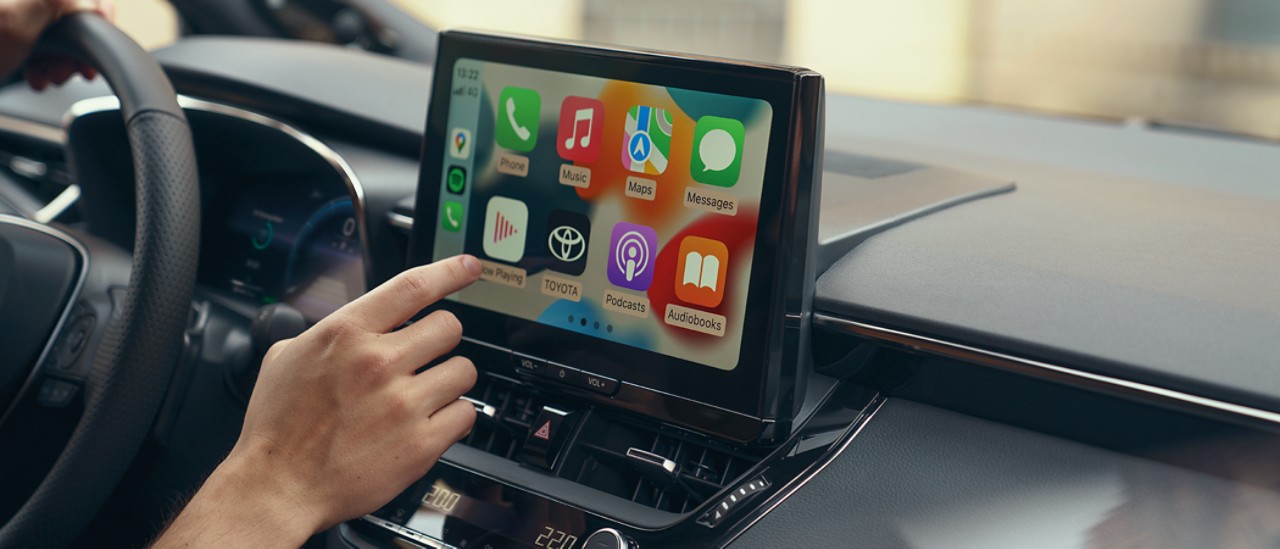 Una persona all’interno di una Toyota interagisce con lo schermo tattile multimediale dell’auto. Sullo schermo è visualizzata la schermata home di Apple CarPlay.