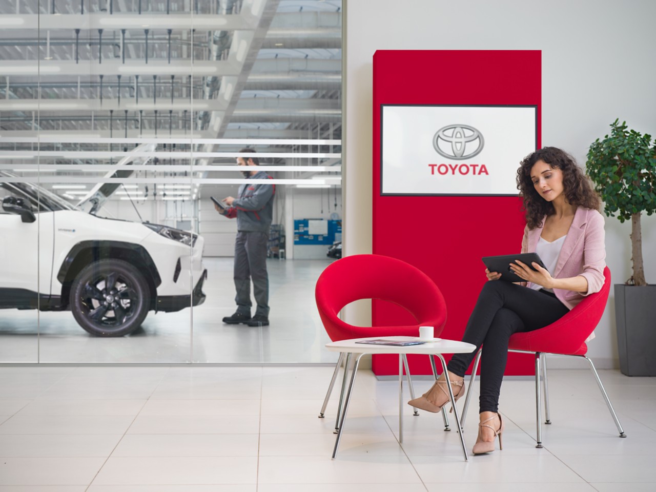 Una donna siede nella sala d’attesa di un’officina Toyota. Sta guardando un tablet, con un logo Toyota visualizzato su uno schermo tv davanti a lei.