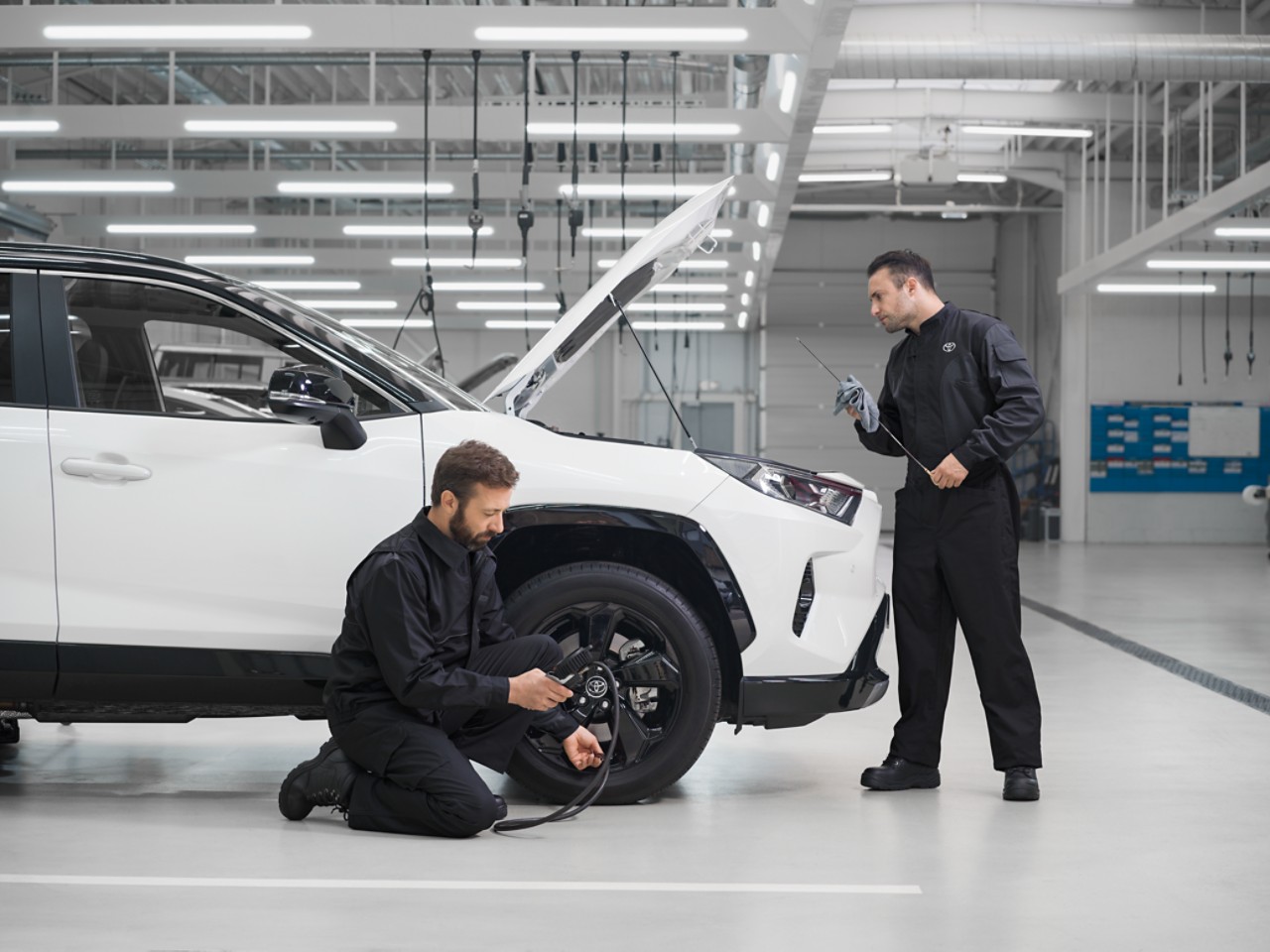 Due tecnici eseguono un intervento di manutenzione su un Toyota RAV4 bianco con il cofano alzato. Uno dei tecnici è in ginocchio e controlla la pressione degli pneumatici, l'altro controlla il livello dell’olio.