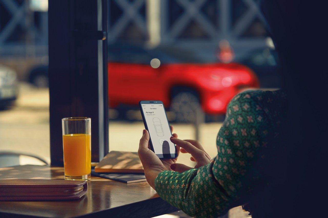 Donna seduta in un bar. Usa l’app TOYOTA MyT sul suo telefono; sullo sfondo si vede la sua Toyota Yaris Cross parcheggiata.