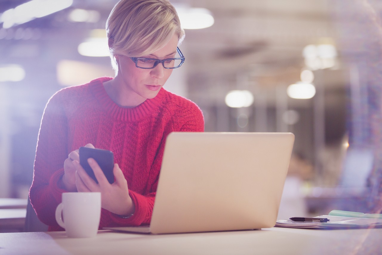 Una donna che indossa un paio di occhiali e un maglione rosso guarda il suo laptop con lo smartphone in mano. Sul tavolo c’è una tazza di caffè.