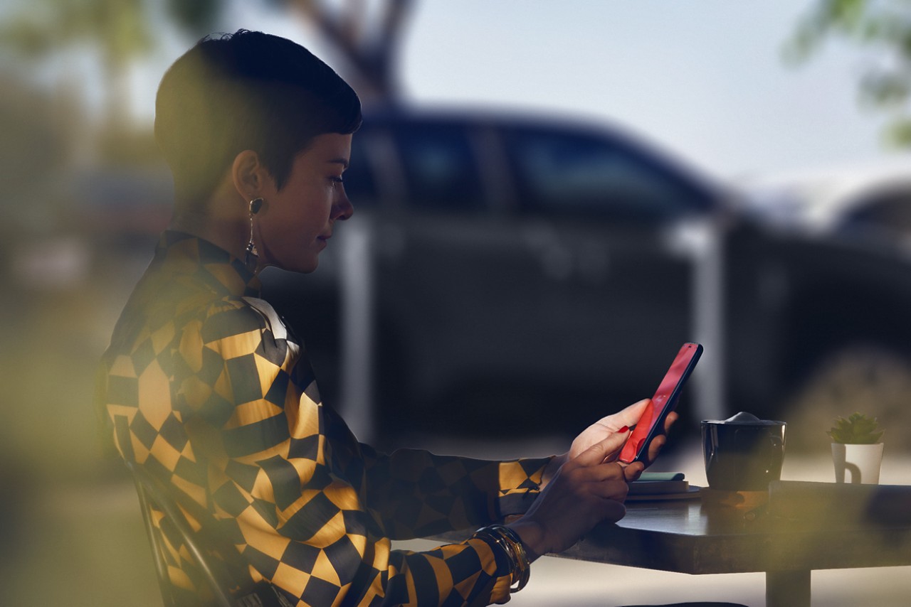 Una signora seduta al tavolo di un ristorante usa l’app MyToyota sul suo telefono; sullo sfondo si vede la sua Toyota Yaris Cross parcheggiata.